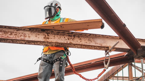 a construction worker wearing personal protective equipment (PPE), such as a helmet, face shield, safety harness, and an orange high-visibility vest to practice safety